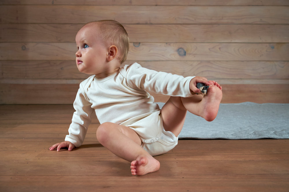 blonde baby sitting on the floor of a wooden room wearing an off-whitebodysuit with snaps at the crotch front. The baby is holding her foot up.