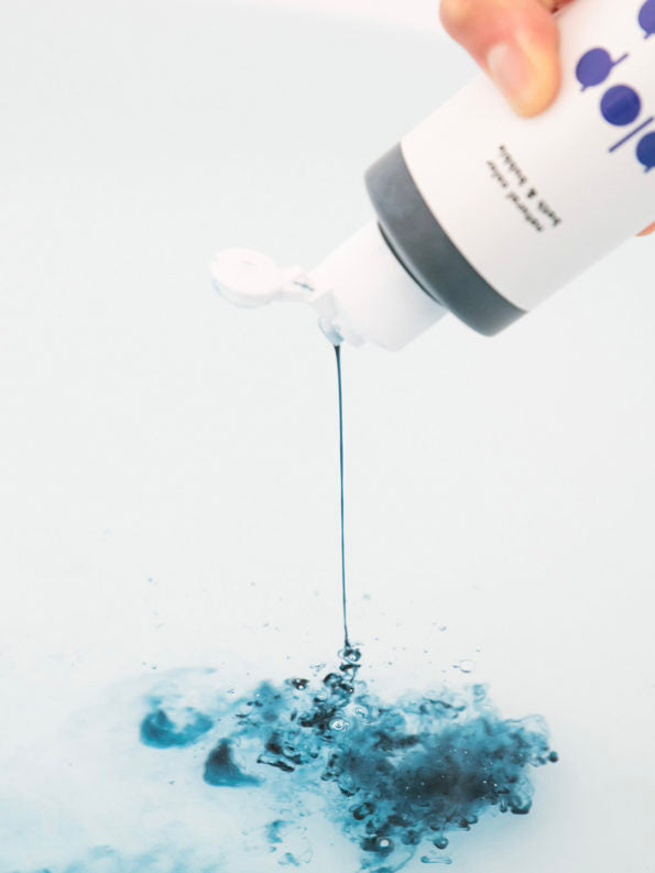 hand pouring a bottle of plop plop colored bubble bath by nahthing project in ocean blue in the water